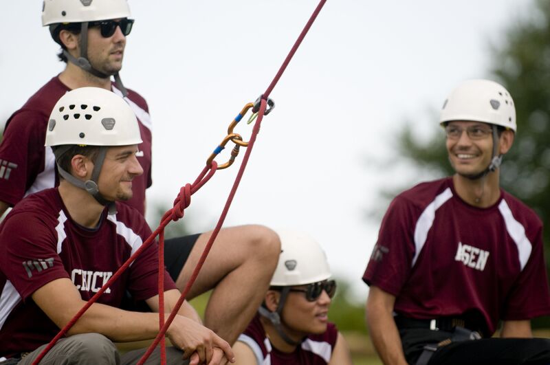 The image shows a group of people wearing helmets and sunglasses, likely preparing for or participating in an outdoor activity like climbing or rappelling. They are dressed in matching maroon shirts with white accents. The focus is on the equipment, including ropes and carabiners, suggesting a focus on safety and teamwork. The expressions on their faces suggest they are enjoying themselves.
